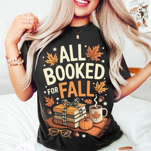 May include: Black t-shirt with the text "ALL BOOKED FOR FALL" in cream and orange. The design includes fall leaves, books, a pumpkin, a mug of coffee, and eyeglasses. A cozy autumn-themed graphic tee.