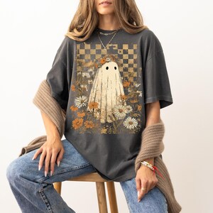Op de afbeelding: Donkergrijs T-shirt met een grillig spookontwerp. Het spook wordt omringd door madeliefjes en oranje bloemen, tegen een geblokte achtergrond. Het shirt heeft een vintage, enigszins vervaagde uitstraling.