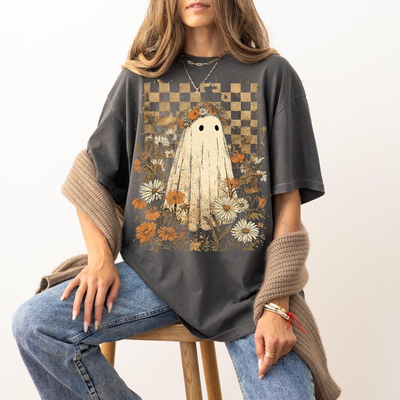 Op de afbeelding: Donkergrijs T-shirt met een grillig spookontwerp. Het spook wordt omringd door madeliefjes en oranje bloemen, tegen een geblokte achtergrond. Het shirt heeft een vintage, enigszins vervaagde uitstraling.