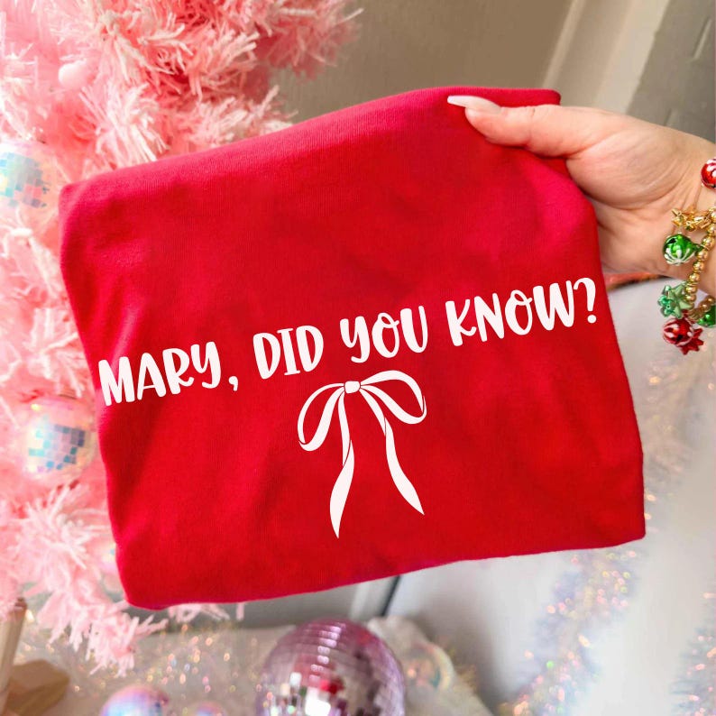 Może przedstawiać: Jasnoczerwona koszulka z białym napisem "MARY, DID YOU KNOW?" oraz białą grafiką kokardy. Koszulka trzymana jest w dłoni. W tle r&oacute;żowa choinka i ozdoby.
