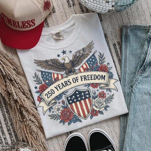 May include: White t-shirt with a patriotic design featuring an eagle, American flags, and the text "250 Years of Freedom." A red and cream baseball cap with the word "TIMELESS" is also visible. The shirt is paired with blue jeans and black sneakers.
