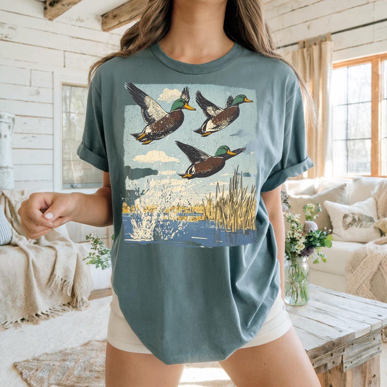May include: Teal-colored t-shirt featuring a graphic of three ducks in flight over a body of water. The ducks are brown, white, and green, with a blue sky and water background. The shirt has short sleeves and a relaxed fit.