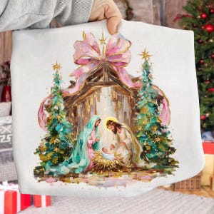May include: A white fabric item featuring a Nativity scene. The artwork depicts Mary, Joseph, and baby Jesus in a manger, surrounded by Christmas trees and a pink bow. The style is painterly with soft colors.
