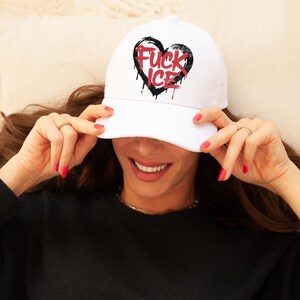 May include: White baseball cap with a black heart graphic and the words "FUCK ICE" in red. The cap has a curved brim and is worn by a person. The text has a dripping effect.