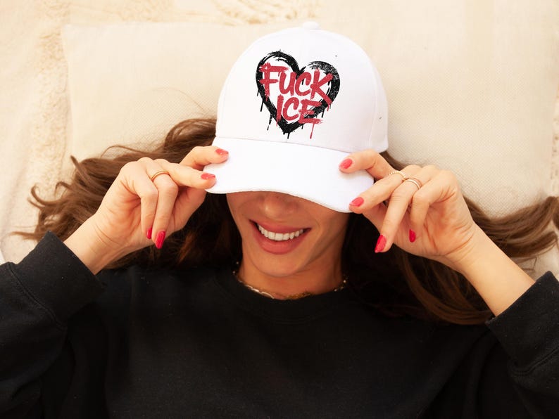 May include: White baseball cap with a black heart graphic and the words "FUCK ICE" in red. The cap has a curved brim and is worn by a person. The text has a dripping effect.