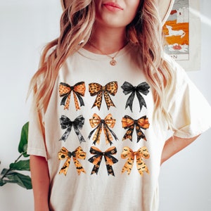 May include: Cream-colored t-shirt featuring a collection of nine decorative bows. The bows showcase various patterns, including leopard print, polka dots, and Halloween-themed designs with bats and pumpkins. The bows are in shades of orange, black, and gold.