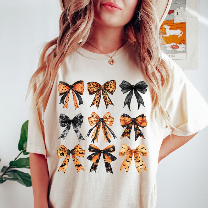 May include: Cream-colored t-shirt featuring a collection of nine decorative bows. The bows showcase various patterns, including leopard print, polka dots, and Halloween-themed designs with bats and pumpkins. The bows are in shades of orange, black, and gold.