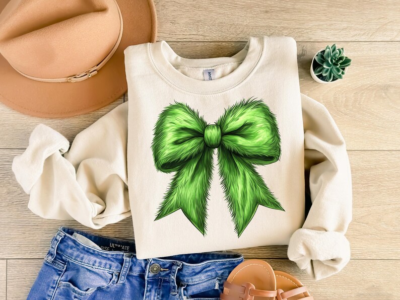 Puede incluir: Sudadera color crema con un gran lazo verde vibrante. El lazo tiene una apariencia esponjosa y texturizada. La sudadera se combina con pantalones cortos de mezclilla azules, un sombrero color canela y sandalias, lo que sugiere un estilo casual y cotidiano.
