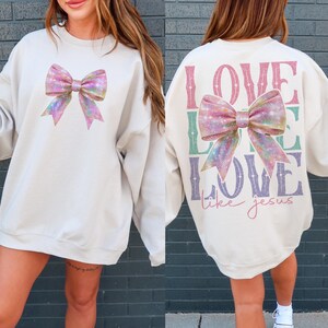 K&ouml;nnte beinhalten: Ein cremefarbener Pullover mit einer glitzernden rosa Schleife auf der Vorderseite und den Worten "LOVE LIKE LOVE like jesus" auf der R&uuml;ckseite.