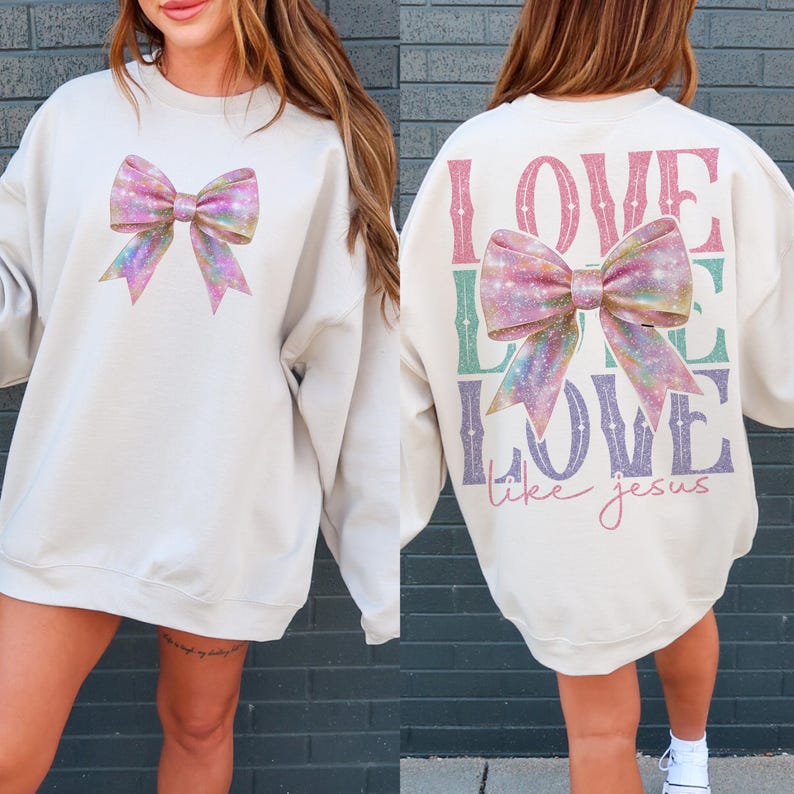 K&ouml;nnte beinhalten: Ein cremefarbener Pullover mit einer glitzernden rosa Schleife auf der Vorderseite und den Worten "LOVE LIKE LOVE like jesus" auf der R&uuml;ckseite.