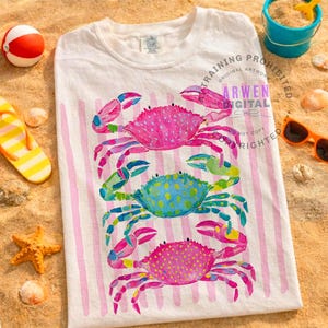 Puede incluir: Camiseta blanca con un dise&ntilde;o de acuarela de tres cangrejos coloridos. Los cangrejos son rosas, azules y rosas, con acentos amarillos y verdes. La camiseta tiene rayas verticales rosas y est&aacute; sobre una superficie arenosa.