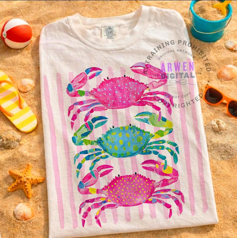 Puede incluir: Camiseta blanca con un dise&ntilde;o de acuarela de tres cangrejos coloridos. Los cangrejos son rosas, azules y rosas, con acentos amarillos y verdes. La camiseta tiene rayas verticales rosas y est&aacute; sobre una superficie arenosa.