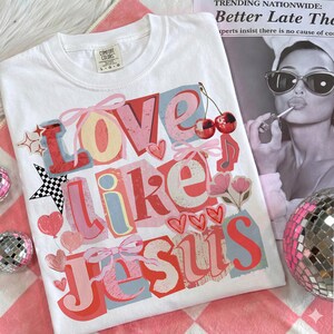 May include: White Comfort Colors t-shirt featuring the text "Love Like Jesus" in a layered, colourful design. The design includes hearts, bows, cherries, and a musical note. The shirt is on a pink and white checkered surface.