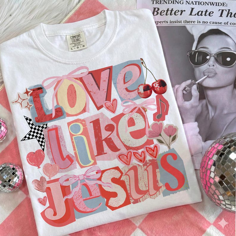 May include: White Comfort Colors t-shirt featuring the text "Love Like Jesus" in a layered, colourful design. The design includes hearts, bows, cherries, and a musical note. The shirt is on a pink and white checkered surface.
