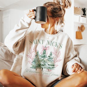 May include: A cream-colored sweatshirt with a graphic that reads "SAINT NICK'S Christmas Tree Farm." The design features three watercolor-style Christmas trees, a pink bow, and leafy accents. The sweatshirt is a cozy, casual style.