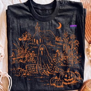 May include: A black t-shirt featuring an orange graphic print of a haunted scene with a church, a crescent moon, corn stalks, pumpkins, and a ghost. A sign in the foreground displays the text "LIVE HERE NOW" in a stylized, weathered font.
