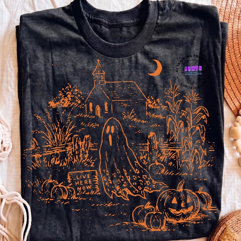 May include: A black t-shirt featuring an orange graphic print of a haunted scene with a church, a crescent moon, corn stalks, pumpkins, and a ghost. A sign in the foreground displays the text "LIVE HERE NOW" in a stylized, weathered font.