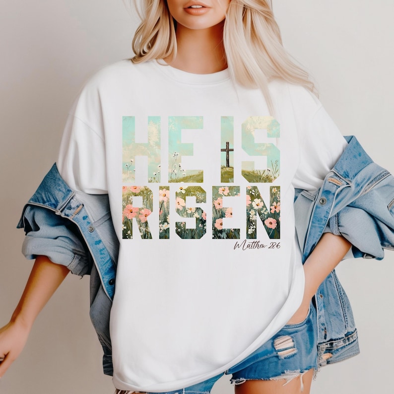 May include: White t-shirt with a floral design that spells out "He is Risen" with a cross and the scripture "Matthew 28:6".