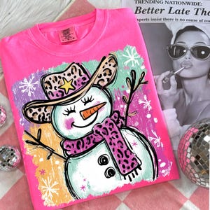 May include: A vibrant pink Comfort Colors sweatshirt with a snowman graphic. The snowman has a leopard print cowboy hat and scarf, a carrot nose, and twig arms. The background includes snowflakes and colourful brushstrokes.