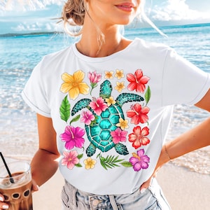 May include: White t-shirt featuring a colourful graphic of a sea turtle surrounded by tropical flowers. The turtle is turquoise blue with a yellow shell and pink, orange, and yellow flowers.