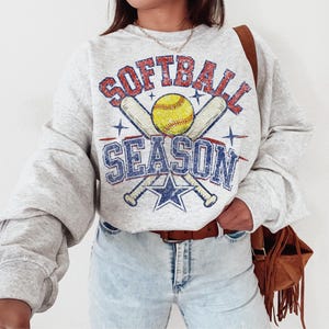 May include: A light gray sweatshirt with the words "SOFTBALL SEASON" in a distressed, vintage style. The design includes a yellow softball, crossed bats, and a blue star. The sweatshirt has long sleeves and a relaxed fit.