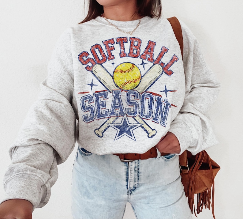May include: A light gray sweatshirt with the words "SOFTBALL SEASON" in a distressed, vintage style. The design includes a yellow softball, crossed bats, and a blue star. The sweatshirt has long sleeves and a relaxed fit.