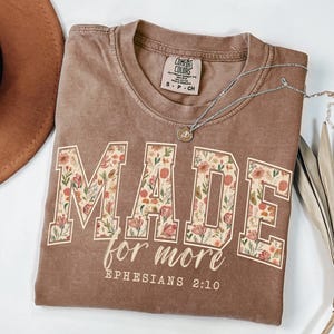 Op de afbeelding: Een bruin t-shirt met de woorden "MADE for more" in een bloemenpatroon. De tekst "Efezi&euml;rs 2:10" staat eronder. Een zilveren ketting met een hanger ligt op het shirt.