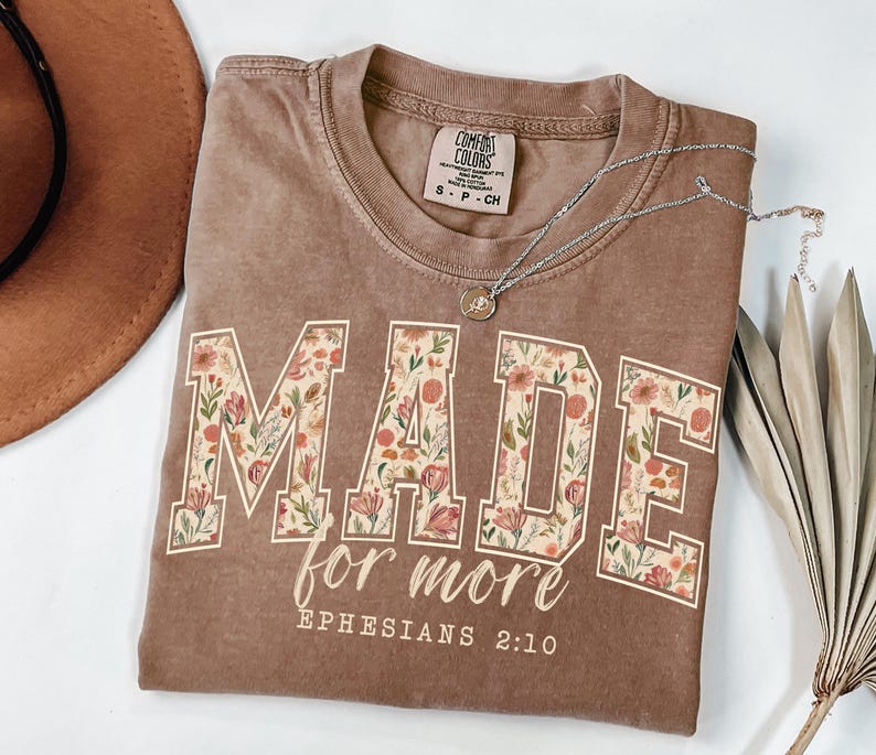 Op de afbeelding: Een bruin t-shirt met de woorden "MADE for more" in een bloemenpatroon. De tekst "Efezi&euml;rs 2:10" staat eronder. Een zilveren ketting met een hanger ligt op het shirt.