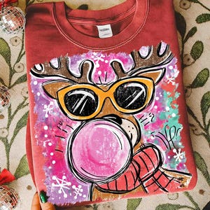 Op de afbeelding: Bordeauxkleurige crewneck sweatshirt met een cartoon rendier met een zonnebril en een kauwgom bel blazend. Het rendier heeft bruine geweien en een rood-zwarte geruite sjaal. Het ontwerp staat tegen een roze en paarse achtergrond met sneeuwvlokken.