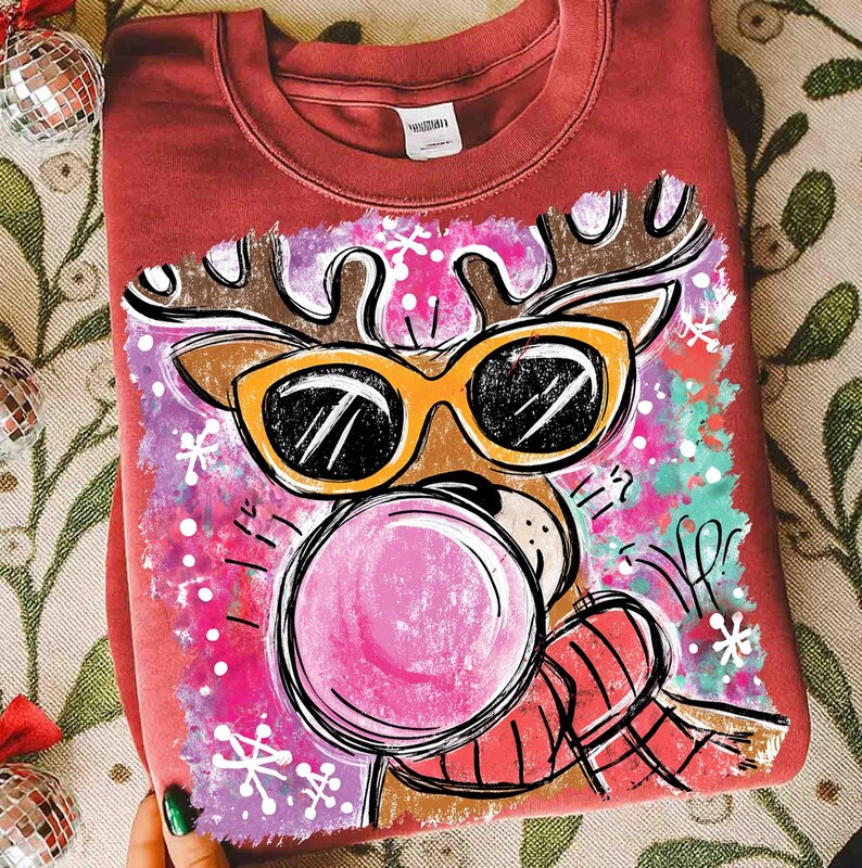Op de afbeelding: Bordeauxkleurige crewneck sweatshirt met een cartoon rendier met een zonnebril en een kauwgom bel blazend. Het rendier heeft bruine geweien en een rood-zwarte geruite sjaal. Het ontwerp staat tegen een roze en paarse achtergrond met sneeuwvlokken.