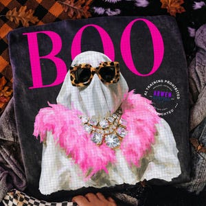 May include: A dark gray t-shirt featuring a white ghost graphic with a grid pattern, wearing leopard print sunglasses and a bright pink feather boa. A large, sparkling rhinestone statement necklace is worn over the boa. The word BOO is printed in large, bold, hot pink letters above the ghost.
