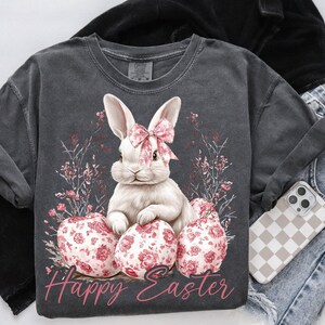 May include: A gray t-shirt with a white bunny rabbit holding pink Easter eggs. The bunny has a pink bow around its neck. The shirt has the text "Happy Easter" printed on it.
