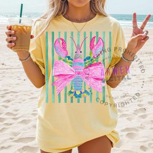 May include: A light yellow t-shirt featuring a colorful lobster design with a large pink bow. The lobster has pink claws and is set against a background of vertical teal stripes. The shirt is worn on a sandy beach.