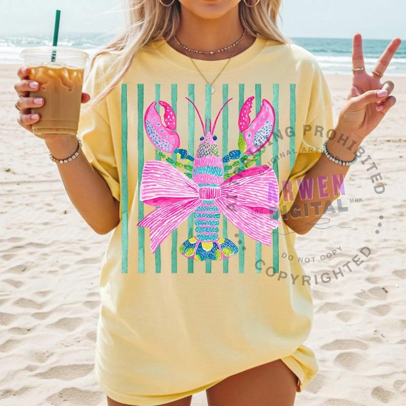 May include: A light yellow t-shirt featuring a colorful lobster design with a large pink bow. The lobster has pink claws and is set against a background of vertical teal stripes. The shirt is worn on a sandy beach.