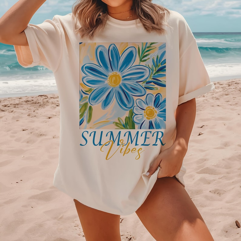 Pu&ograve; includere: Una maglietta bianca con una grafica di fiori blu e foglie verdi. Il testo "SUMMER Vibes" &egrave; stampato in giallo sotto la grafica.