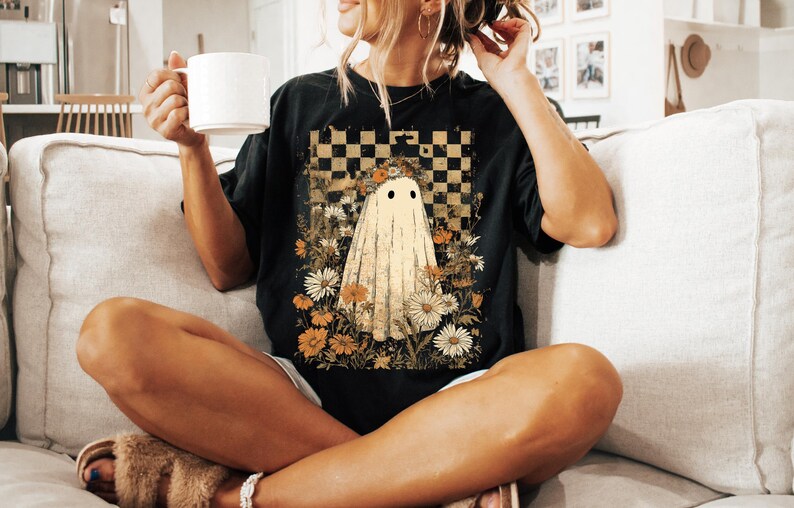 Op de afbeelding: Zwart T-shirt met een spookontwerp tegen een bloemen- en geblokte achtergrond. De geest is cr&egrave;mekleurig met zwarte ogen. De bloemen zijn in tinten geel, oranje en wit. Het shirt heeft een casual, relaxte pasvorm.