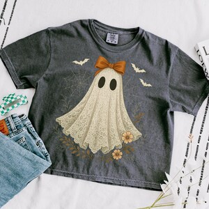 May include: Dark gray t-shirt featuring a ghost design with a lace-like appearance, a brown bow, and bat illustrations. The shirt is paired with blue jeans and a green and white hair clip. A magazine is also visible.