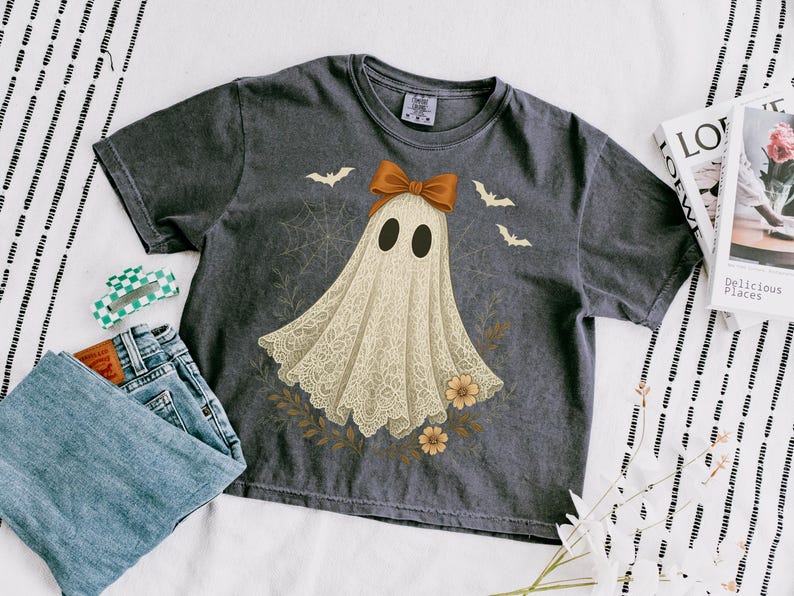 May include: Dark gray t-shirt featuring a ghost design with a lace-like appearance, a brown bow, and bat illustrations. The shirt is paired with blue jeans and a green and white hair clip. A magazine is also visible.