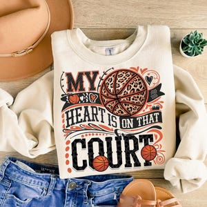 May include: A cream-coloured sweatshirt with the text "My heart is on that court" in black and orange. A basketball with a leopard print design is featured in the centre of the text.
