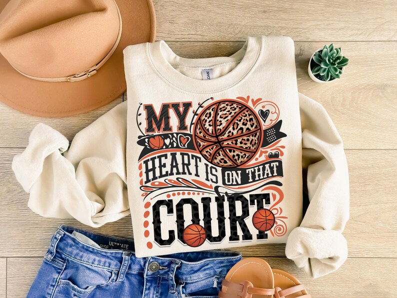 May include: A cream-coloured sweatshirt with the text "My heart is on that court" in black and orange. A basketball with a leopard print design is featured in the centre of the text.