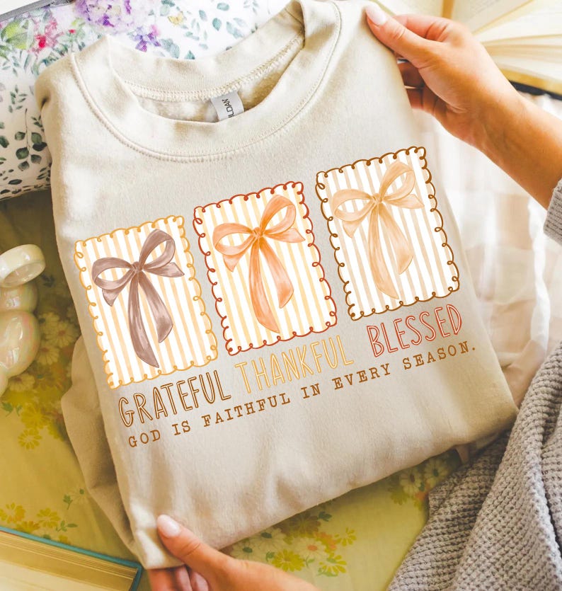 May include: A beige sweatshirt featuring three rectangular designs with bows in shades of brown and orange. The text "Grateful Thankful Blessed" is printed below the designs, with the phrase "God is faithful in every season."