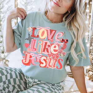 May include: A pale green t-shirt featuring the text "Love Like Jesus" in a layered, colourful design. The graphic includes hearts, cherries, and a star with a chequered pattern. The shirt is part of a casual clothing collection.