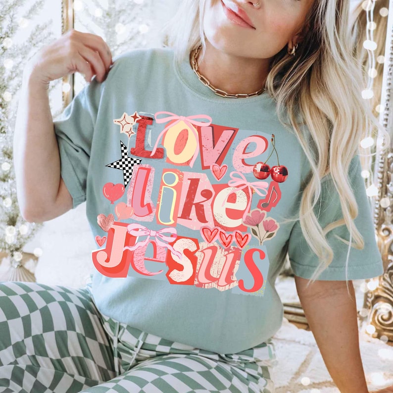 May include: A pale green t-shirt featuring the text "Love Like Jesus" in a layered, colourful design. The graphic includes hearts, cherries, and a star with a chequered pattern. The shirt is part of a casual clothing collection.