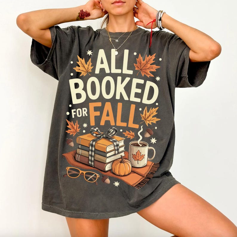 May include: Dark gray t-shirt with the text "ALL BOOKED FOR FALL" and fall-themed graphics. The design includes books, a pumpkin, a coffee mug, and autumn leaves. The shirt is a relaxed fit.
