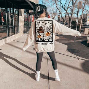 May include: A cream-coloured sweatshirt with a Halloween graphic on the back. The design includes a haunted house, pumpkins, bats, and the text "Halloweentown Est. 1998". The sweatshirt is worn with black leggings and white trainers.