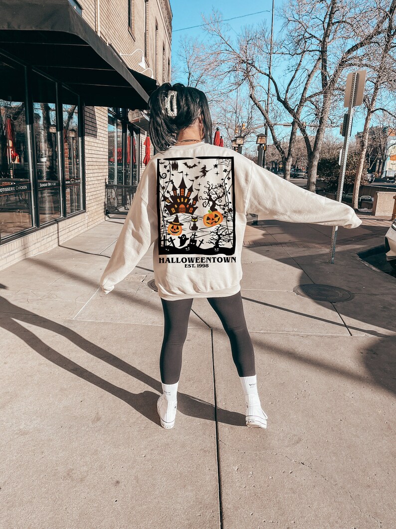 May include: A cream-coloured sweatshirt with a Halloween graphic on the back. The design includes a haunted house, pumpkins, bats, and the text "Halloweentown Est. 1998". The sweatshirt is worn with black leggings and white trainers.