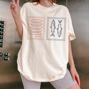May include: Cream-colored t-shirt featuring two postage stamp designs. One stamp shows five baguettes, the word "forever" below. The other stamp has two fish. The shirt is worn by a person.