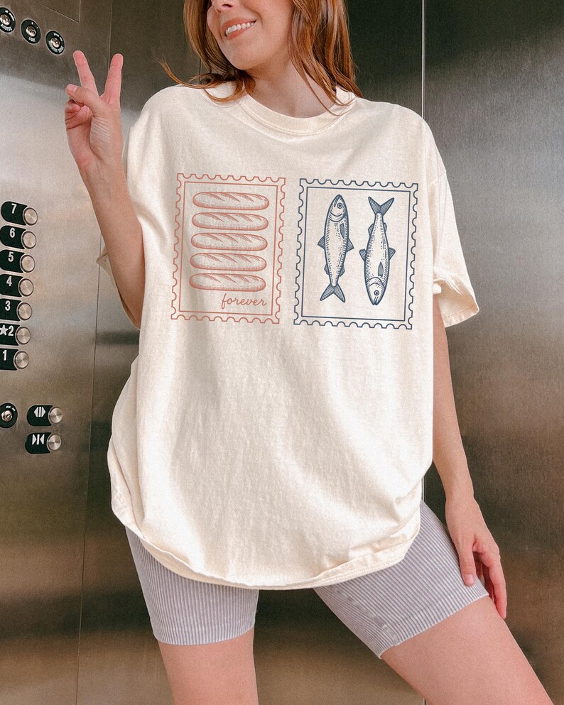 May include: Cream-colored t-shirt featuring two postage stamp designs. One stamp shows five baguettes, the word "forever" below. The other stamp has two fish. The shirt is worn by a person.