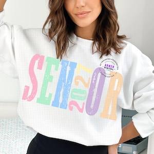 Puede incluir: Una sudadera blanca de manga larga con un fondo de patr&oacute;n de cuadr&iacute;cula y la palabra SENIOR en letras grandes, desgastadas y de colores pastel. El n&uacute;mero 2027 est&aacute; impreso en el centro en colores amarillo, rosa y verde pastel.