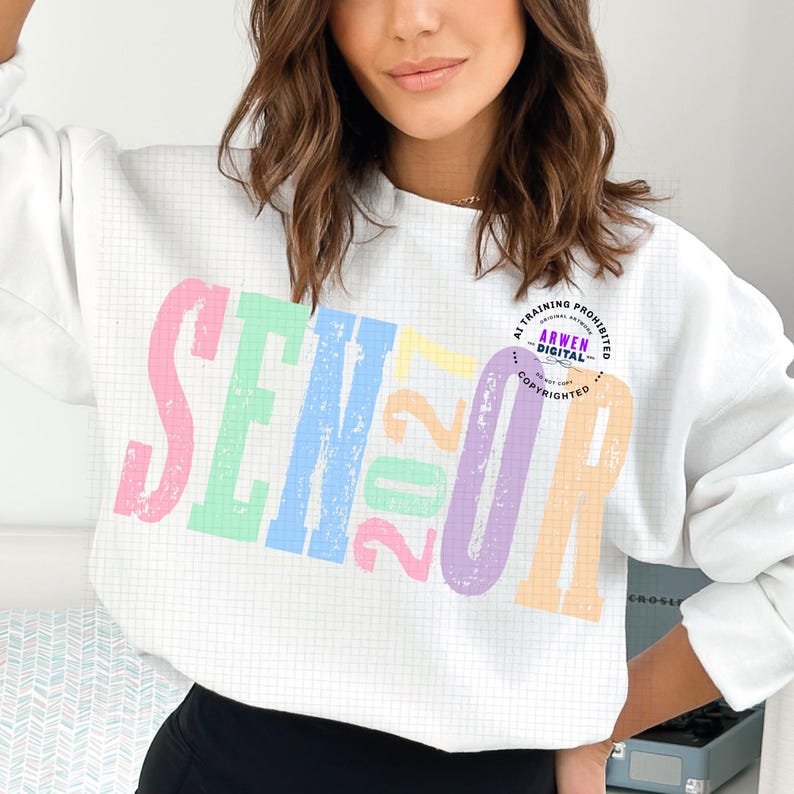 Puede incluir: Una sudadera blanca de manga larga con un fondo de patr&oacute;n de cuadr&iacute;cula y la palabra SENIOR en letras grandes, desgastadas y de colores pastel. El n&uacute;mero 2027 est&aacute; impreso en el centro en colores amarillo, rosa y verde pastel.